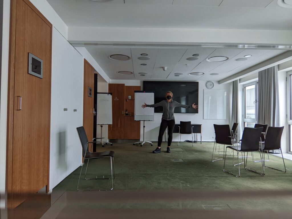 The conference room I thought I would have. I'm standing with my arms outstretched. The room is mainly empty but for a couple clusters of chairs and a few easels.