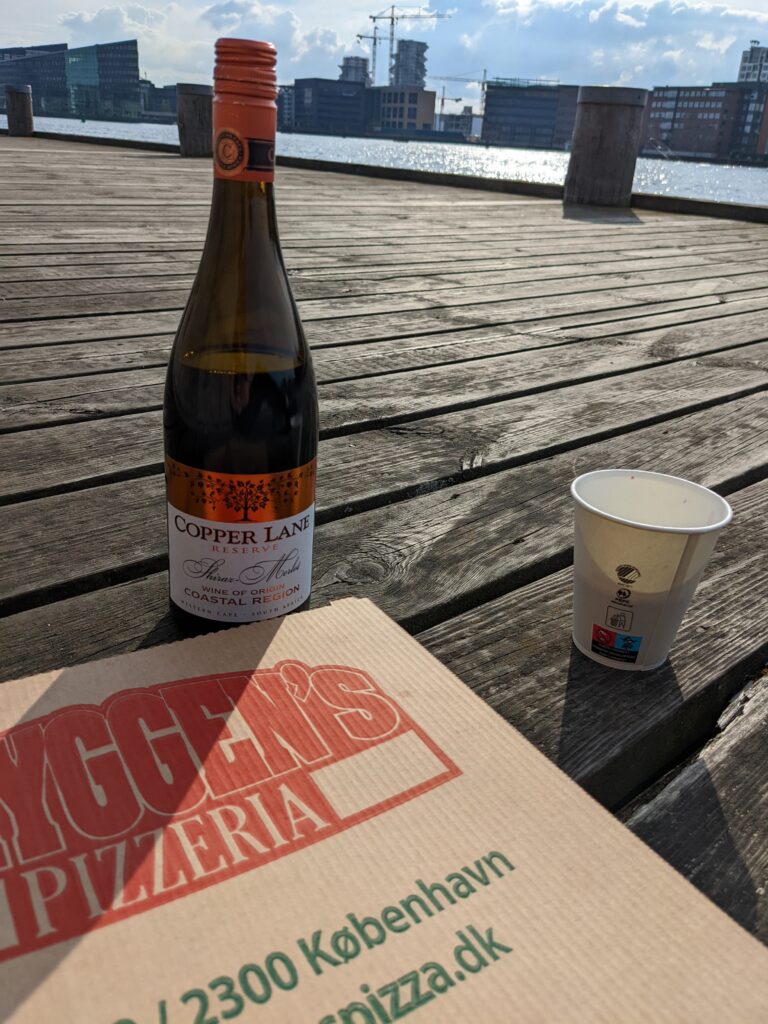 Cardboard pizza box, bottle of red wine, and paper cup on dock by a canal.